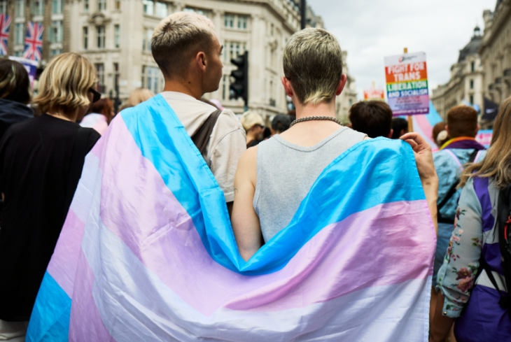 تم تصوير شخصين من الخلف، واقفين معًا وعلم Trans Pride ملفوف حولهما