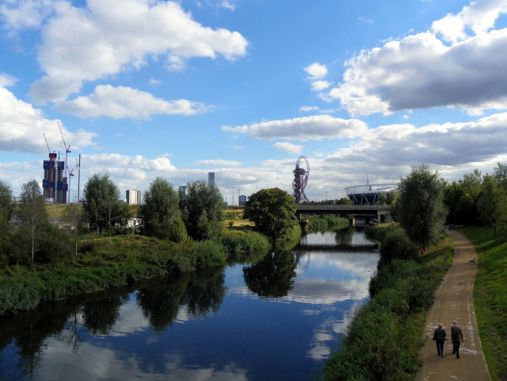 A scenic view of a river reflecting a blue sky with white clouds, with a walking path on the right and the London Olympic Park skyline, including the ArcelorMittal Orbit and Olympic Stadium, in the distance.