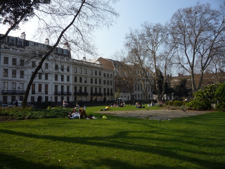 People relaxing on a sunny day in a grassy park square, surrounded by tall white townhouses and bare trees.