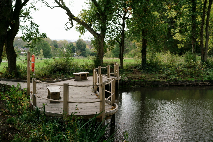 A curved wooden boardwalk with rope railings extends over a calm pond, featuring two small stone benches and an orange lifebuoy nearby, surrounded by lush green trees and a grassy field.