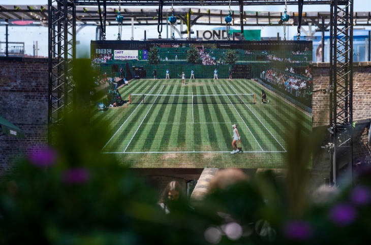 Free screenings of Wimbledon 2026 matches in London: a view through a bush of a large screen showing a tennis match