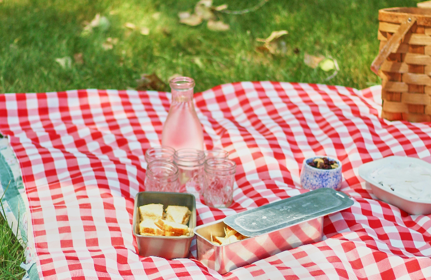 A red and white checkered picnic blanket spread on green grass, topped with a glass carafe, small jars, metal food containers with sandwiches, a small bowl of berries, and a white dish. A woven picnic basket sits in the background.