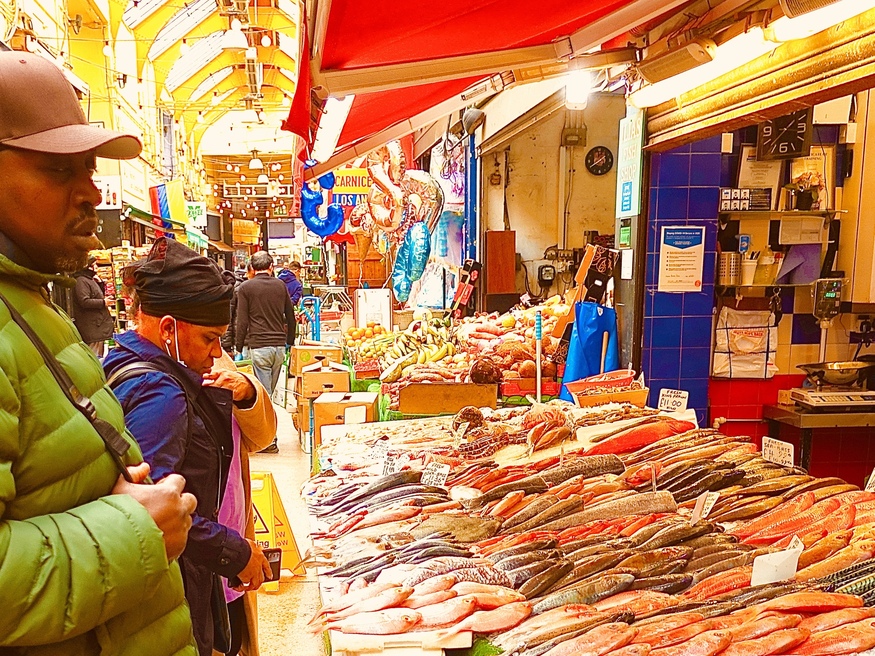 A man in a green puffer jacket and a woman in a blue coat look at a large display of fresh fish at an outdoor market stall under a red awning.