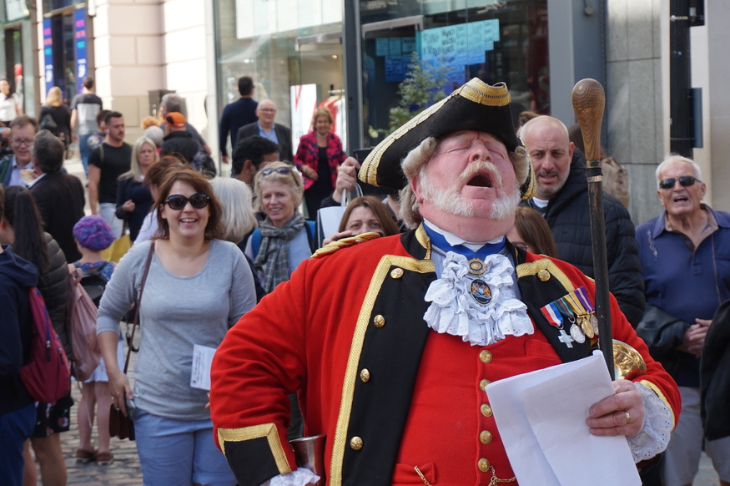 A Town Crier in red tunic and hat bellowing loudly surrounded by onlookers