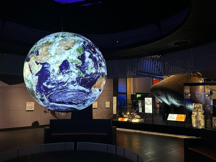 Free things to do in London in the 2026 summer holidays:  A large, glowing globe of Earth suspended in a dark museum exhibit, with a space capsule and informational displays in the background.