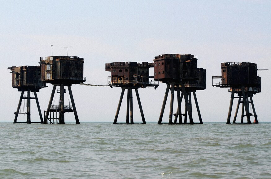 Things to do in London today: A series of rusted, derelict Maunsell sea forts standing on stilts in the middle of a calm ocean under a pale sky.
