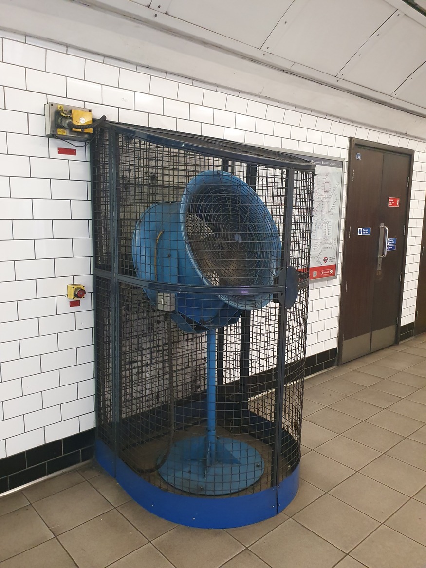 A large blue industrial fan stands inside a tall, curved metal cage against a white-tiled wall in a subway station.