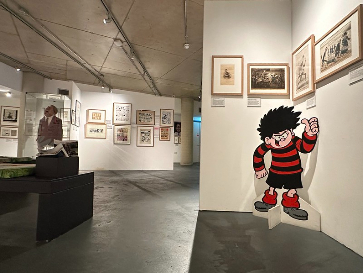 What's on in London this weekend: A wide-angle view of a cartoon and comic art exhibition. In the foreground, a large cardboard cutout of Dennis the Menace stands against a white wall. The gallery features numerous framed comic strips and illustrations on the walls, with a glass display case and a black bench visible in the background.