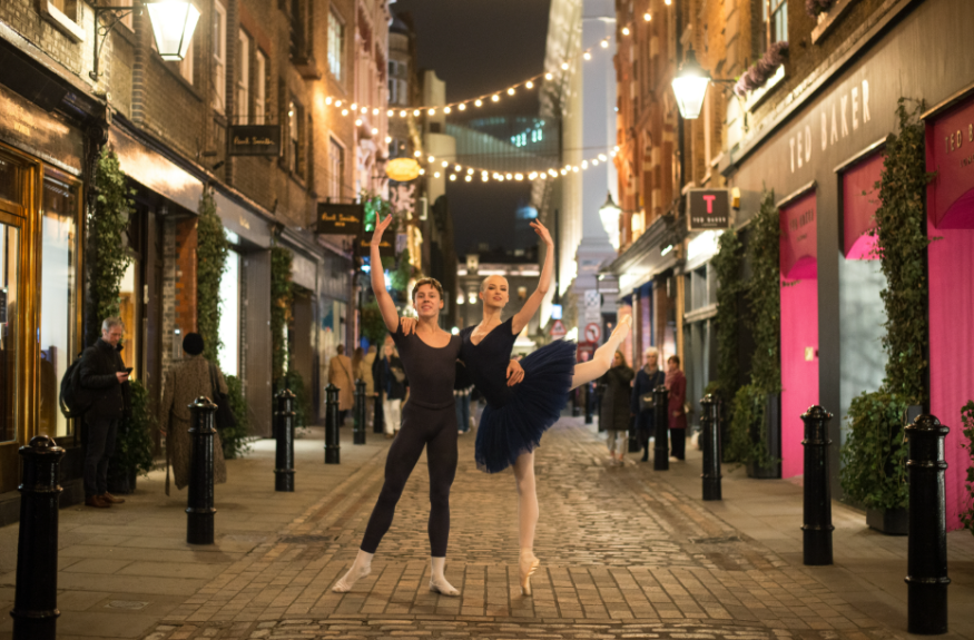 Top events in London today: Two ballerinas posing for a photo down an alleyway lined with shops, beneath fairy lights