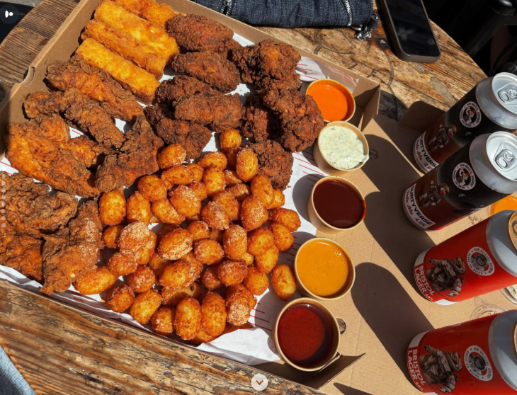 A tray of food including chicken wings and tater tots, alongside pots of dip and cans of beer