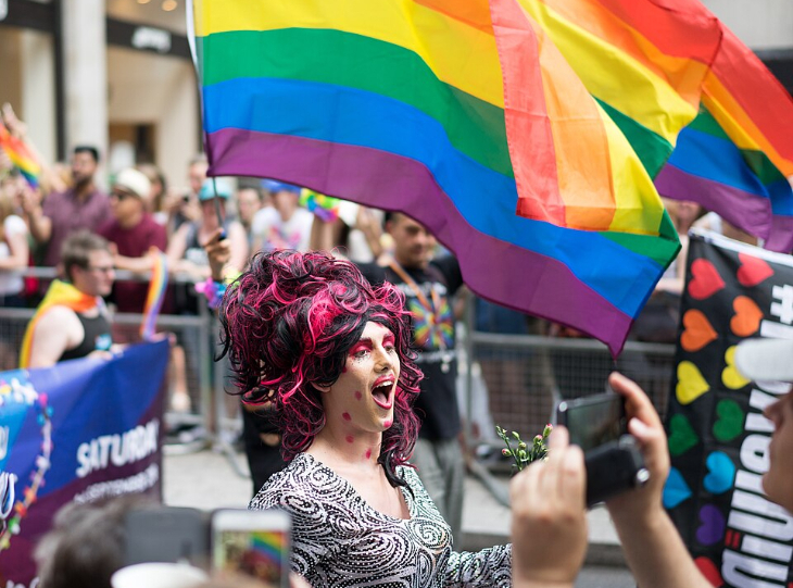 Top events in London today: A person in drag with voluminous pink and black hair and a patterned silver top smiles at a pride parade as a large rainbow flag waves above them and onlookers take photos.