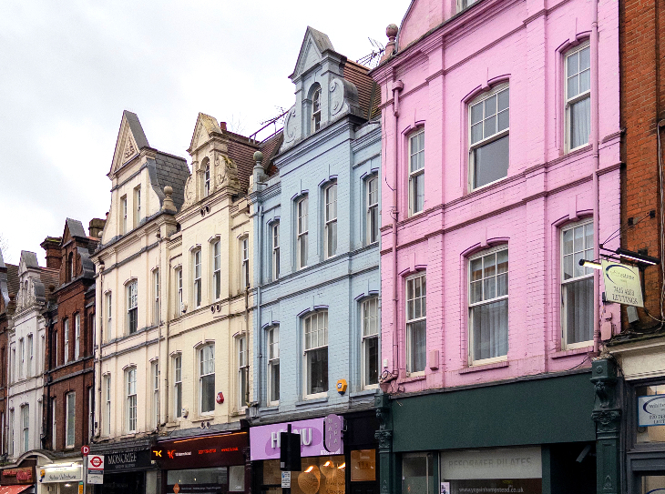 Weekend events in London: A row of colorful, narrow Victorian-style buildings with ornate gables in pink, blue, and cream stand above ground-floor shops on a city street. 