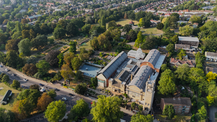 Top events in London this weekend: High-angle aerial view of the Horniman Museum and Gardens in London, featuring the historic clock tower, Victorian conservatory, and modern barrel-roofed galleries surrounded by lush green trees and a residential neighborhood.