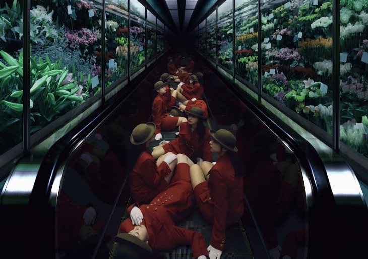 Colour photograph of several women
wearing a red blazer-skirt uniform and brown hats sitting
along a moving walkway.