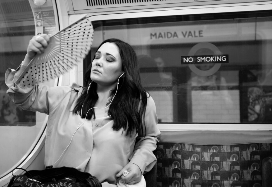 A black and white photo of a woman sitting on a London Underground train, eyes closed, using a hand fan to cool herself. The station sign for Maida Vale is visible through the window behind her.