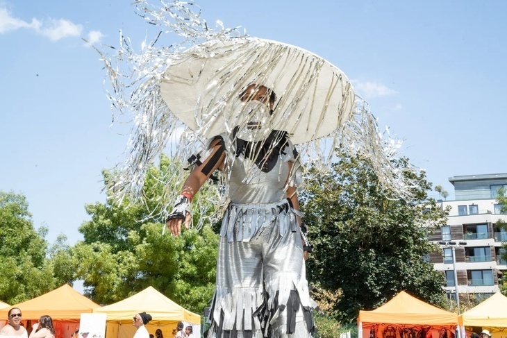 A person in a very large hat is partly obscured by the silvery ribbons dangling thereof while he/she/they dance around in a Kentish Town park