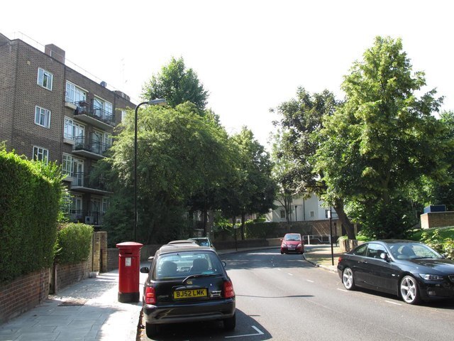 A quiet residential street in London featuring a red post box on the sidewalk, parked cars, and a brick apartment building surrounded by lush green trees.