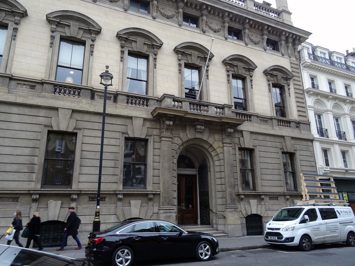 A grand, multi-story stone building with ornate window carvings and a central arched entrance, situated on a city street with pedestrians, a black car, and a white van parked in front.