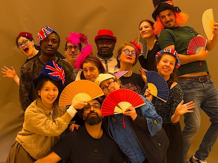 A group of actors with fans (of the wafting type) against a mustard-coloured background. 