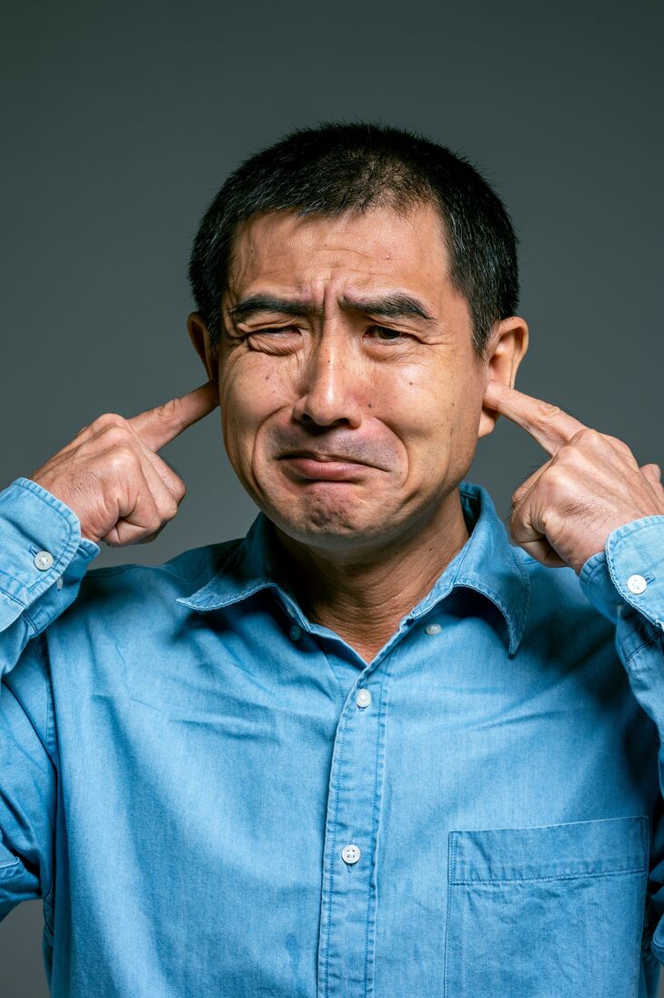 A middle-aged man in a blue button-down shirt winces and plugs his ears with his index fingers against a gray background.