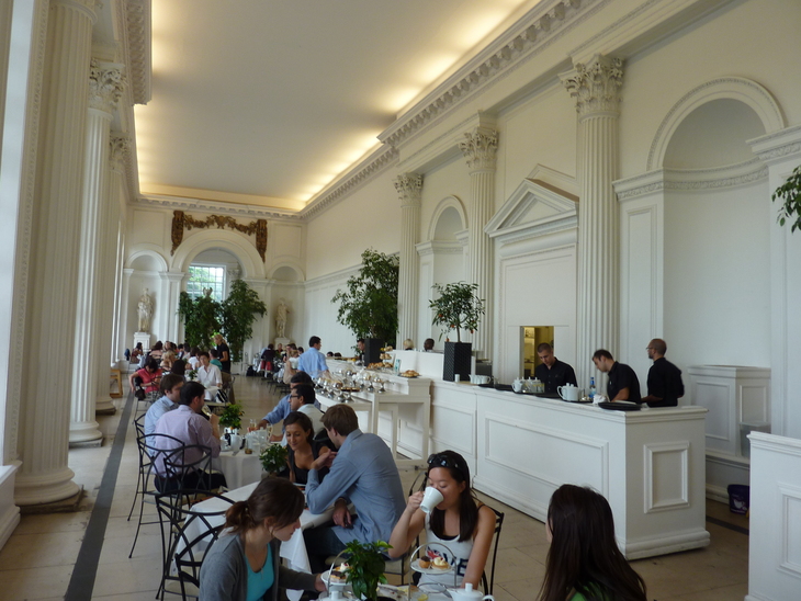 Where to have afternoon tea in London: People sit at tables in a grand, white hall with high ceilings and Corinthian columns, while staff work behind a long white counter.