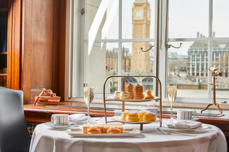 Top afternoon tea in London:  a table laid with a white cloth, with an afternoon tea stand. The River Thames and the Elizabeth Tower are visible through the window in the background.