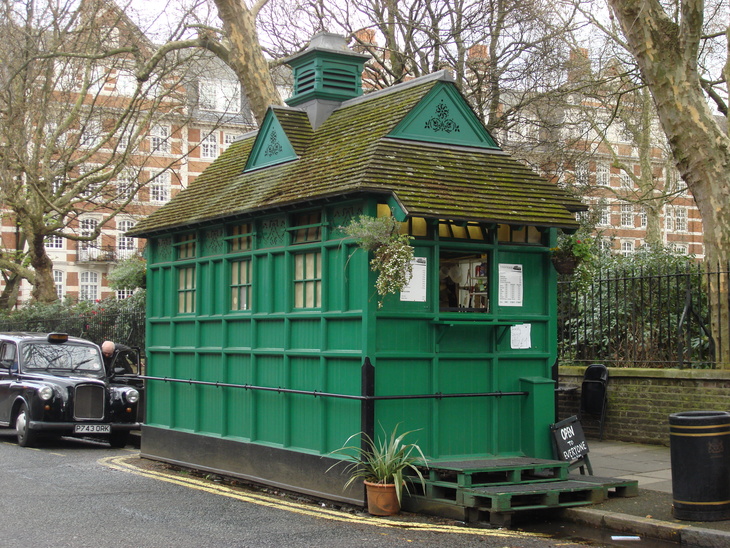 A small, green wooden hut with a mossy shingled roof and decorative gables stands on a London street next to a black taxi. The hut features a service window with menus and is surrounded by bare trees and brick buildings.