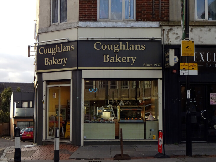 A street-level view of Coughlans Bakery, a corner shop with a black and gold sign that reads "Since 1937." The storefront features large glass windows displaying baked goods and an open doorway on the left.