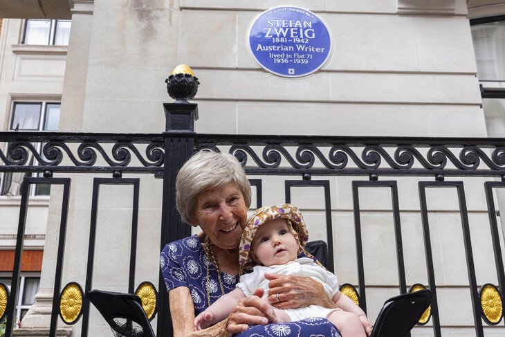 An older woman in front of the plaque, with a baby