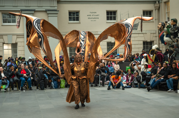 Someone wearing a large bronze Carnival-style costume in front of a crowd at the National Maritime Museum