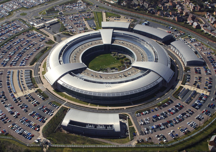 Aerial view of the Government Communications Headquarters (GCHQ) in Cheltenham, England, featuring a large, circular, ring-shaped building with a central courtyard, surrounded by extensive parking lots and residential areas.