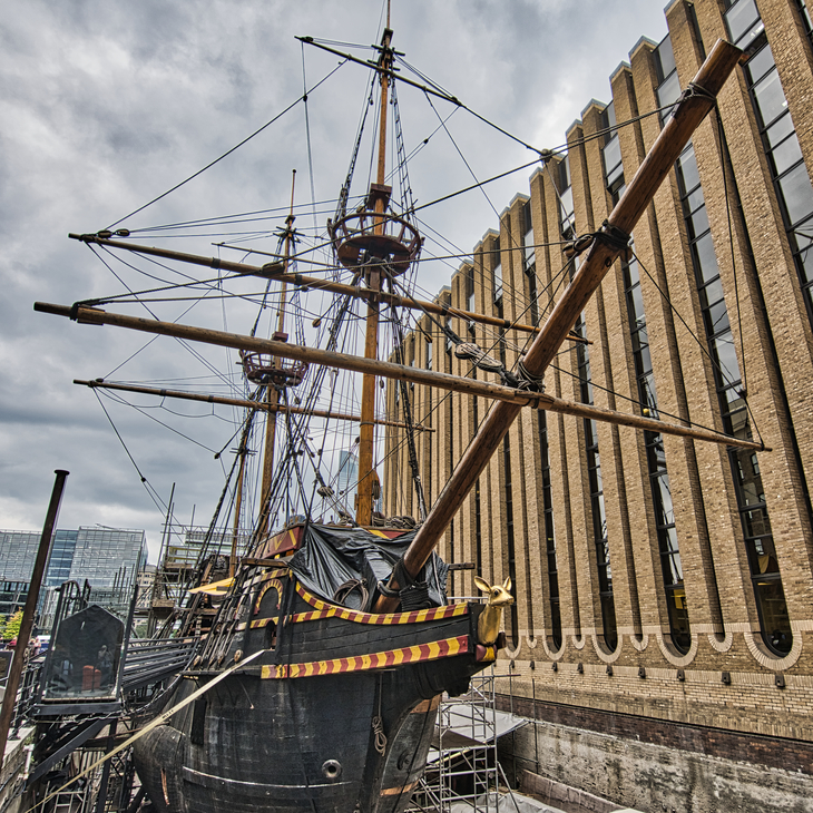 A low-angle shot of the Golden Hinde, a full-scale replica of the 16th-century galleon, docked in London next to a modern brick building under a cloudy sky.
