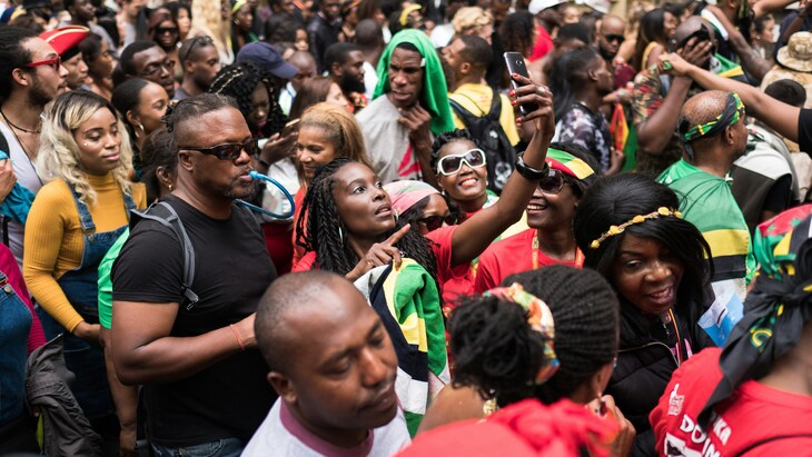 A large, diverse crowd of people gathered closely together at an outdoor event, many wearing vibrant colors and flags. In the center, a woman holds up a smartphone to take a photo or video, while others around her smile, talk, and look toward the same direction.