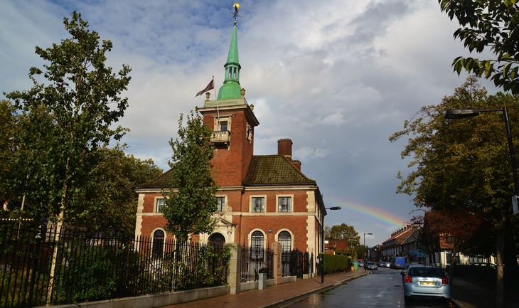 A red brick building with a green spire and a Norwegian flag stands under a cloudy sky with a rainbow in the background.