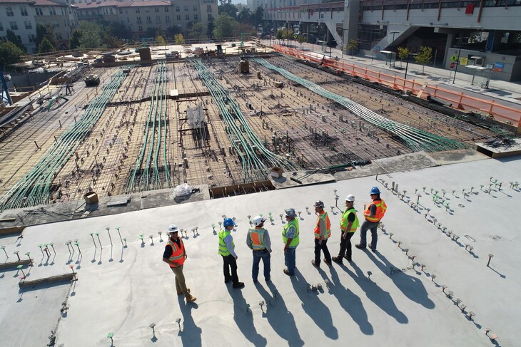 Seven construction workers in safety vests and hard hats stand on a concrete slab overlooking a large construction site with rebar and green post-tensioning cables.