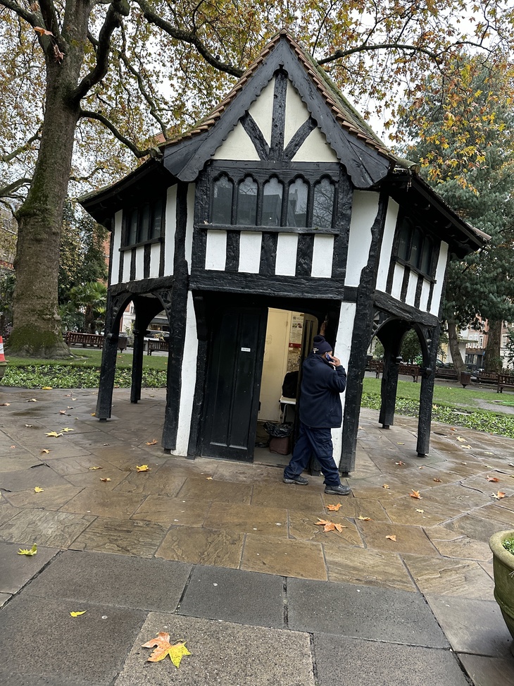 A small, two-story Tudor-style timber-framed building with a gabled roof stands in a paved park area. A person in a dark jacket and blue pants walks through an open doorway on the ground floor.