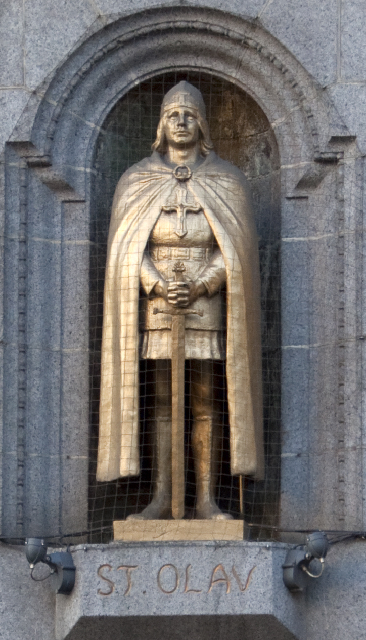 A golden statue of St. Olav stands in a stone niche, wearing a helmet, cloak, and armor with a cross on the chest, holding a sword pointed downward.
