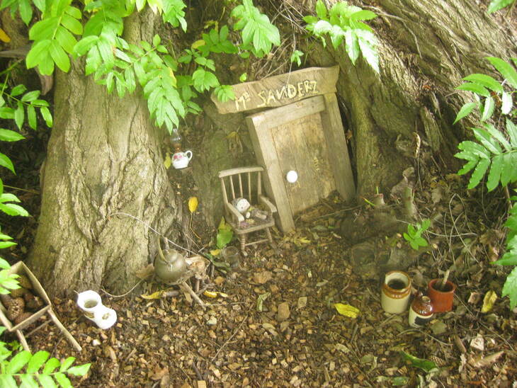 A miniature fairy house built into the base of a large tree, featuring a wooden door with a sign reading "MR SANDERZ," a tiny chair, a wheelbarrow, and various small ceramic pots and kettles.