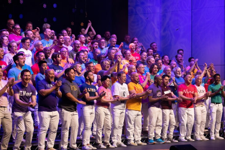 A choir comprised of men wearing white trousers and different coloured t shirts