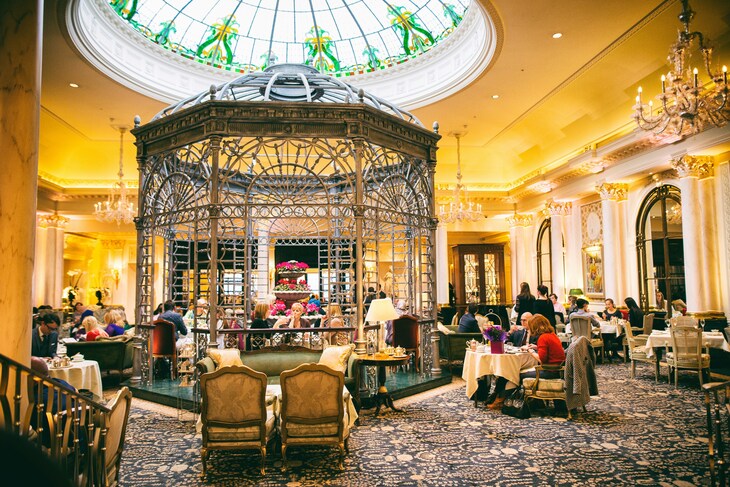 London's finest afternoon tea: Elegant tea room with a large ornate metal gazebo in the center, set under a stained-glass dome ceiling and crystal chandeliers. Patrons sit at tables and on plush sofas throughout the room, which features patterned carpets and classical architectural details.