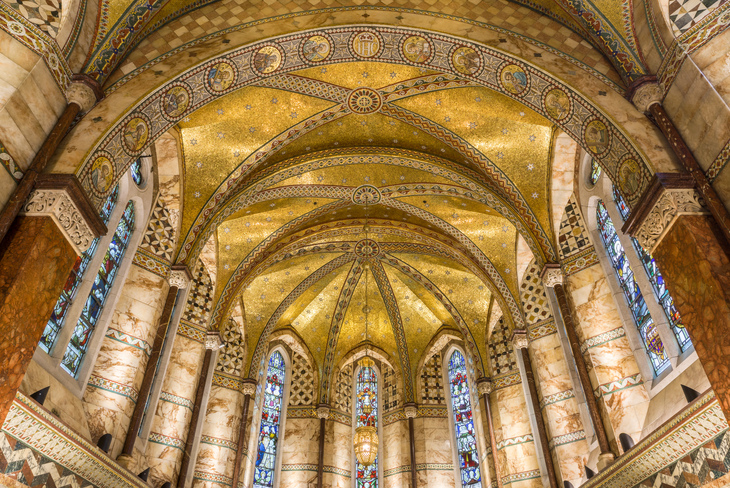 Low-angle view of the ornate, gold-tiled vaulted ceiling and marble walls of the Fitzrovia Chapel in London, featuring stained glass windows and intricate mosaic patterns.