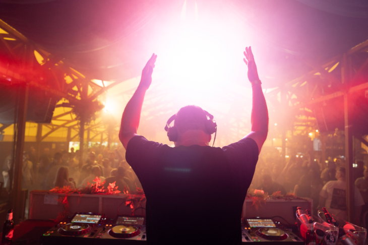  A back view of a DJ silhouetted with his arms in the air, playing to a crowd