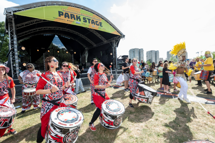 Drummers and a performer in a carnival-style costume performing on grass in front of a festival stage