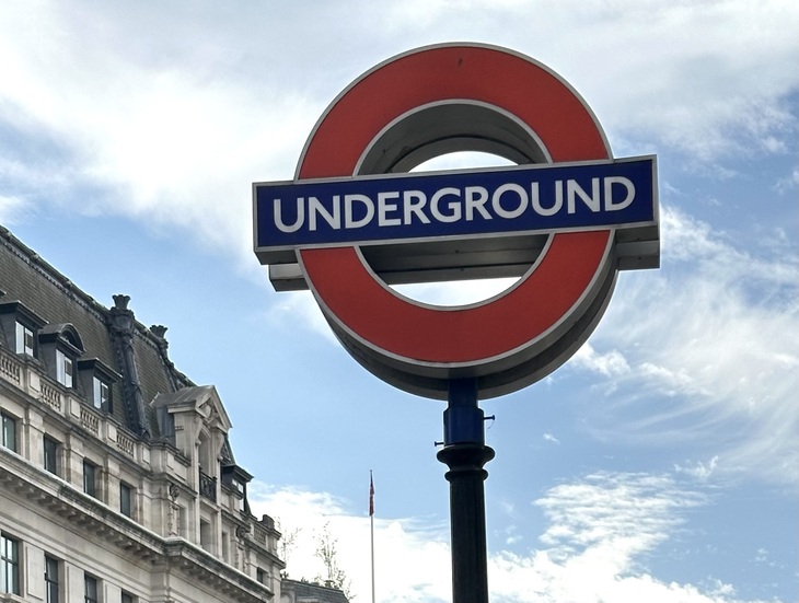 A London Underground roundel sign on a black post against a blue sky with wispy clouds and a historic stone building in the background.