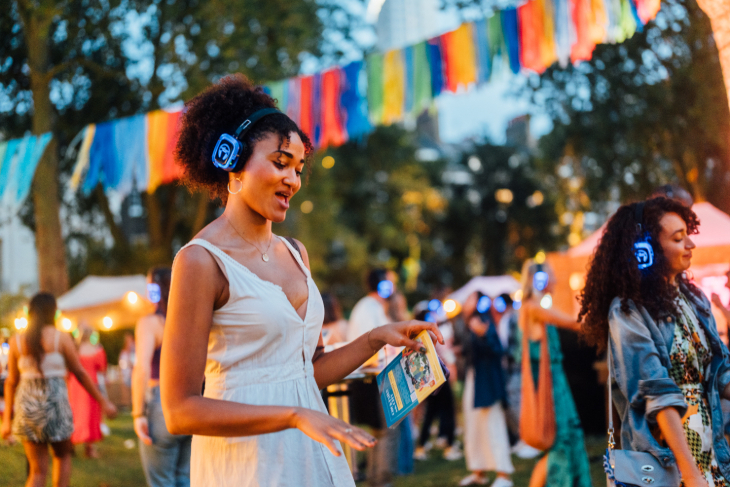 A woman wearing a white dress and headphones, dancing at an open-air silent disco.