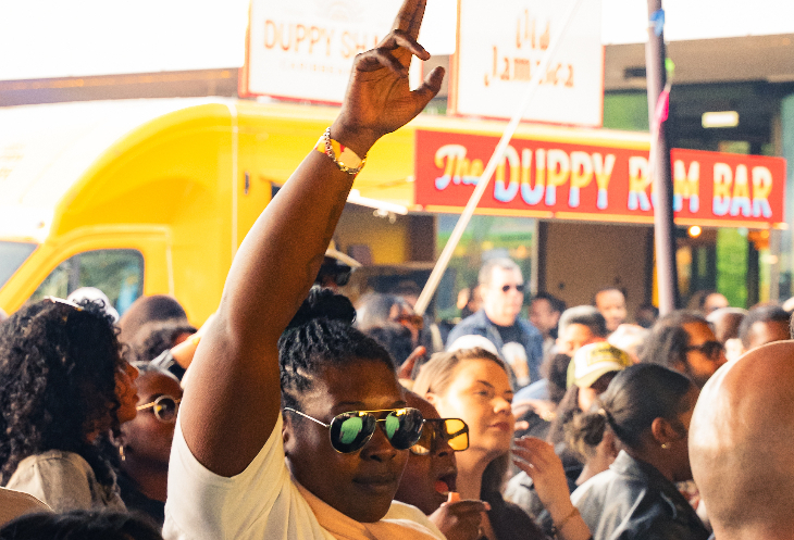 A woman with her arm raised in the air in a crowd at a festival, with a rum bar in the background