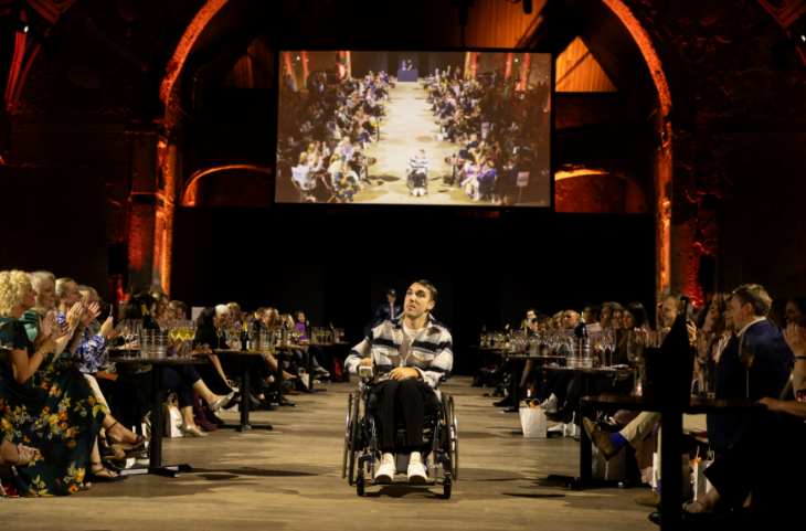 September 2026 top events in London: A person in a wheelchair on a catwalk-style aisle with people watching from tables on either side. Unlike a traditional catwalk, this catwalk is on the floor, at the same level as the audience