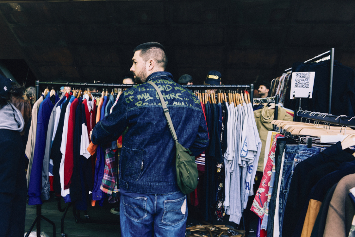 A person in denim jacket and jeans standing in front of a rack of clothes at a flea market