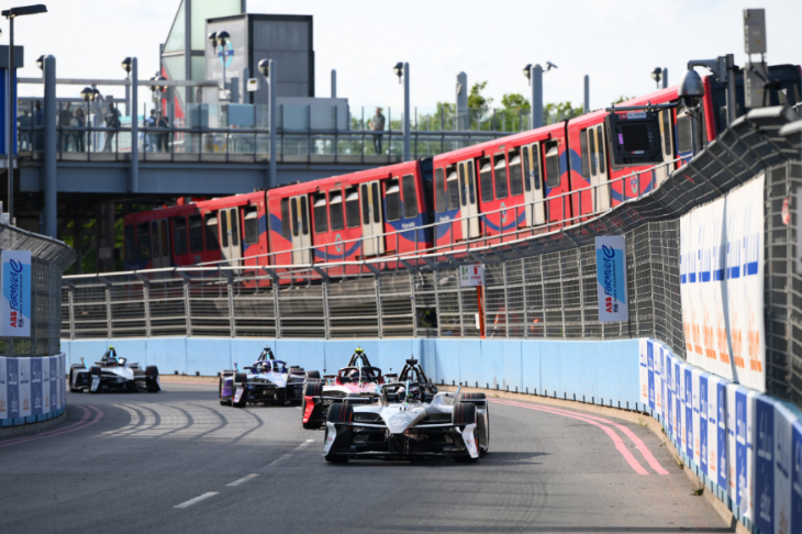 Motorsports cars racing along a road, with a red DLR train passing by overhead