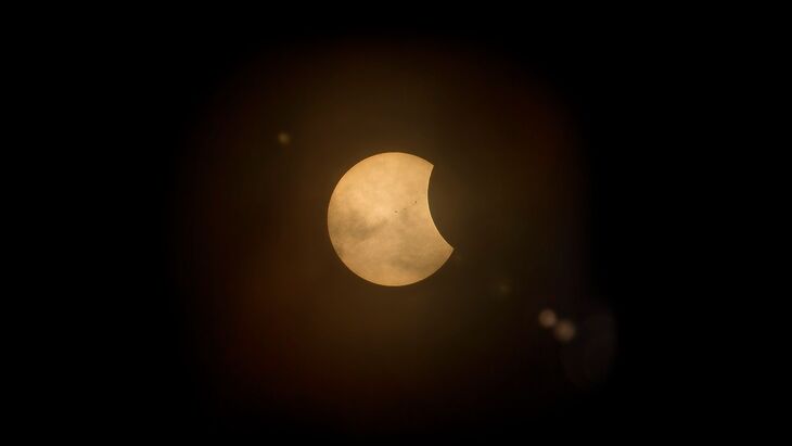 A partial solar eclipse appears as a glowing orange crescent against a dark sky, with faint lens flares and wispy clouds visible across the sun's surface.